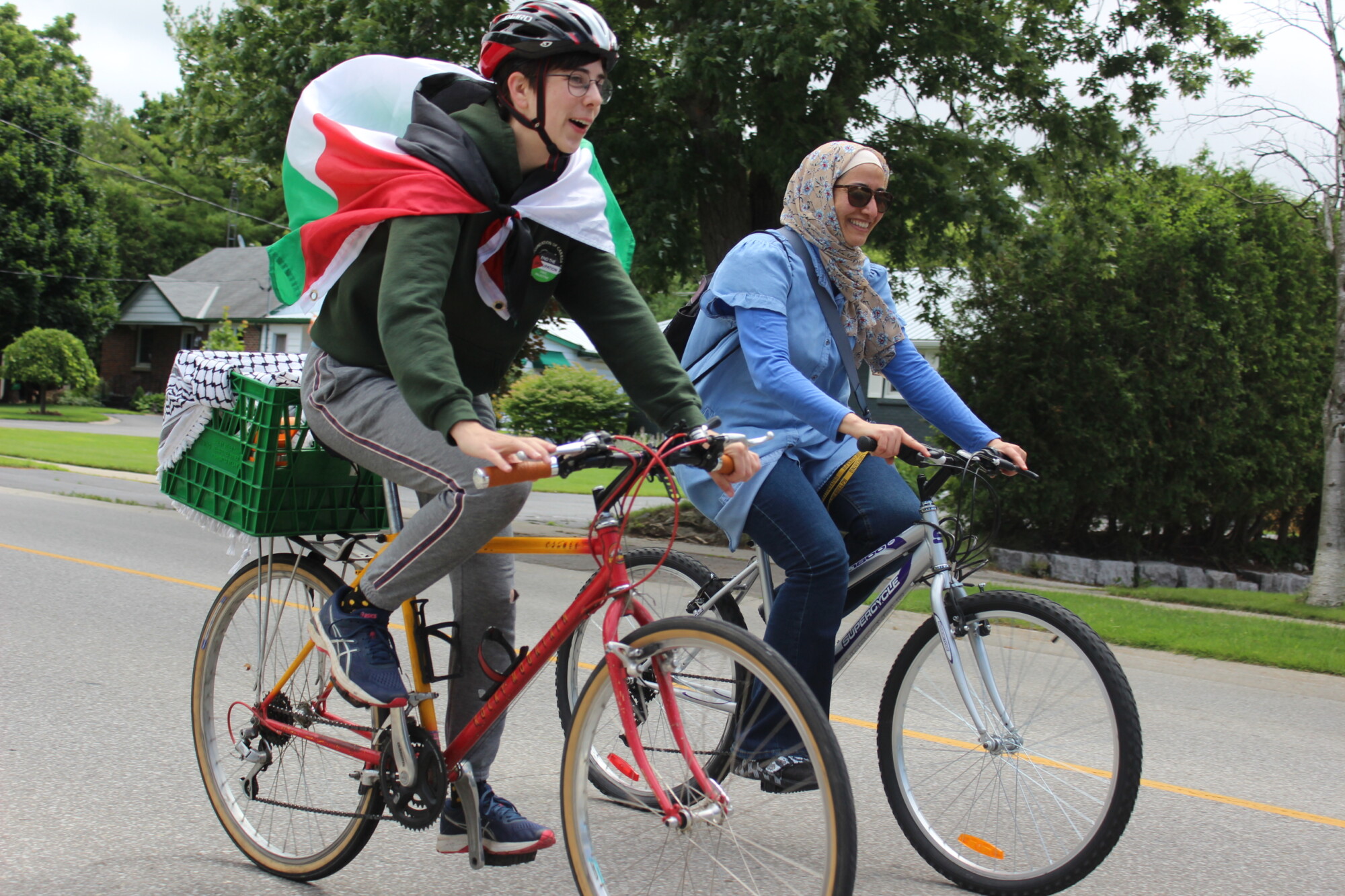 Photograph of two participant cyclists, one wearing a Palestinian flag around their shoulders, riding their bikes in the Waterloo Region Ride for Palestine, an event run by Interfaith Justice on June 30th 2024.