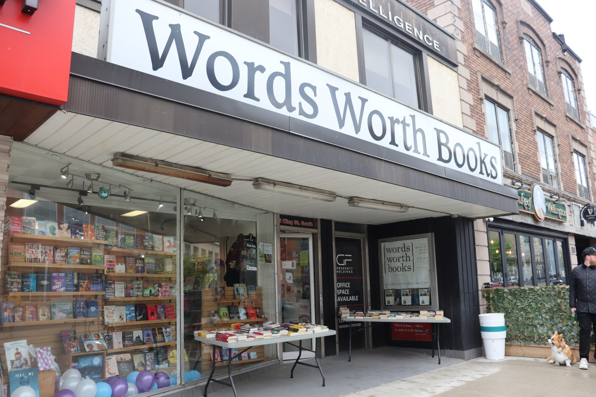 Photograph of the front door and main sign of Words Worth Bookstore, located on King Street in Uptown Waterloo, Ontario. There are tables set up beside the main entrance, covered in books, and the windows are filled with books on shelves and balloons celebrating the store's 40th anniversary on May 11th, 2024.