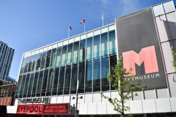 Photograph taken on a sunny day of the front of THEMUSEUM in Kitchener, Ontario. A red and black sign hangs under the windows advertising the current exhibit.