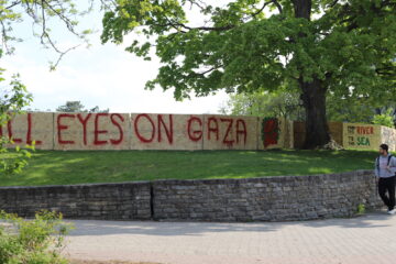 Photo of the plywood wall used as the UW Occupy encampment boarders, on the campus of the University of Waterloo in Ontario, Canada. The message spray painted in red on the plywood reads "All Eyes on Gaza".