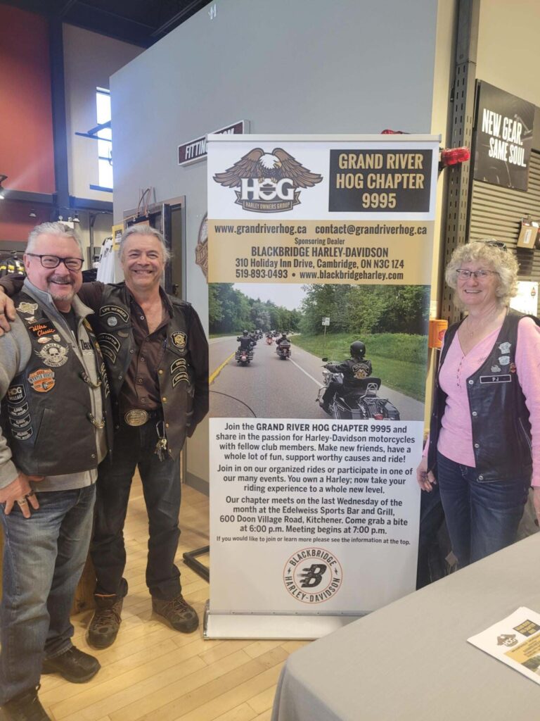 Photo taken during the Blackbridge Harley-Davidson dealership's International Women's Day Event "Inspire Inclusion" of three attendees standing next to a poster for the Grand Rive Hog Chapter 9995.