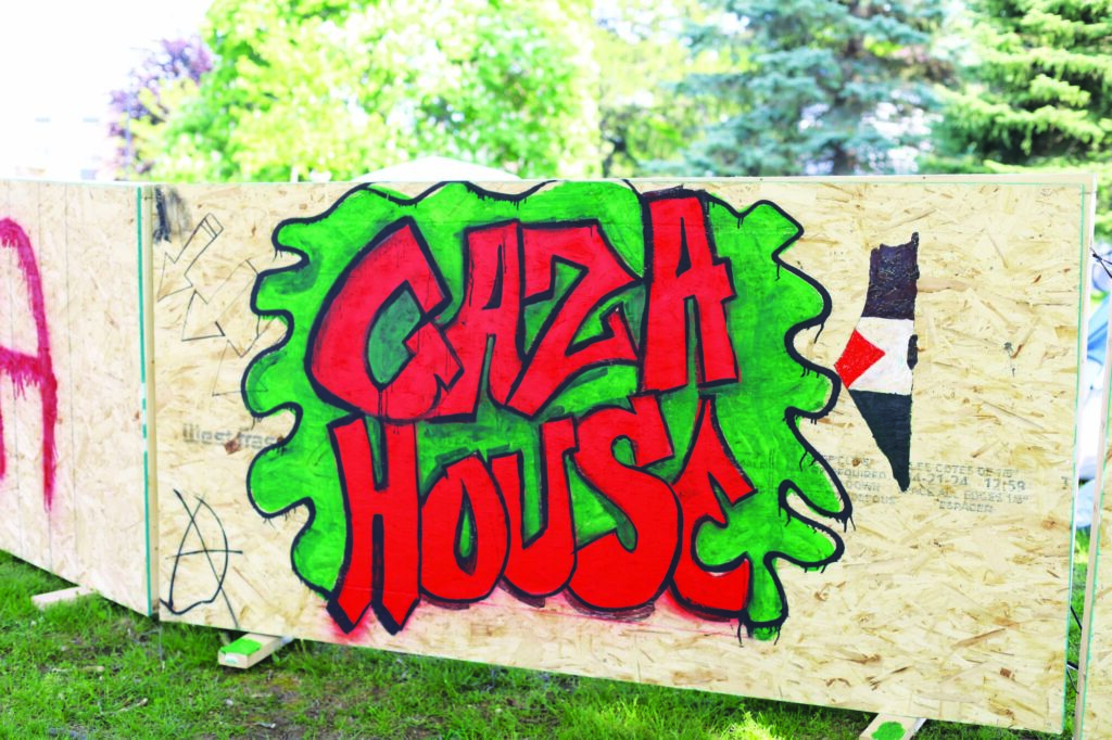 Photograph of a sign painted in bright red and green on the side of a sheet of particle board reading "GAZA HOUSE".