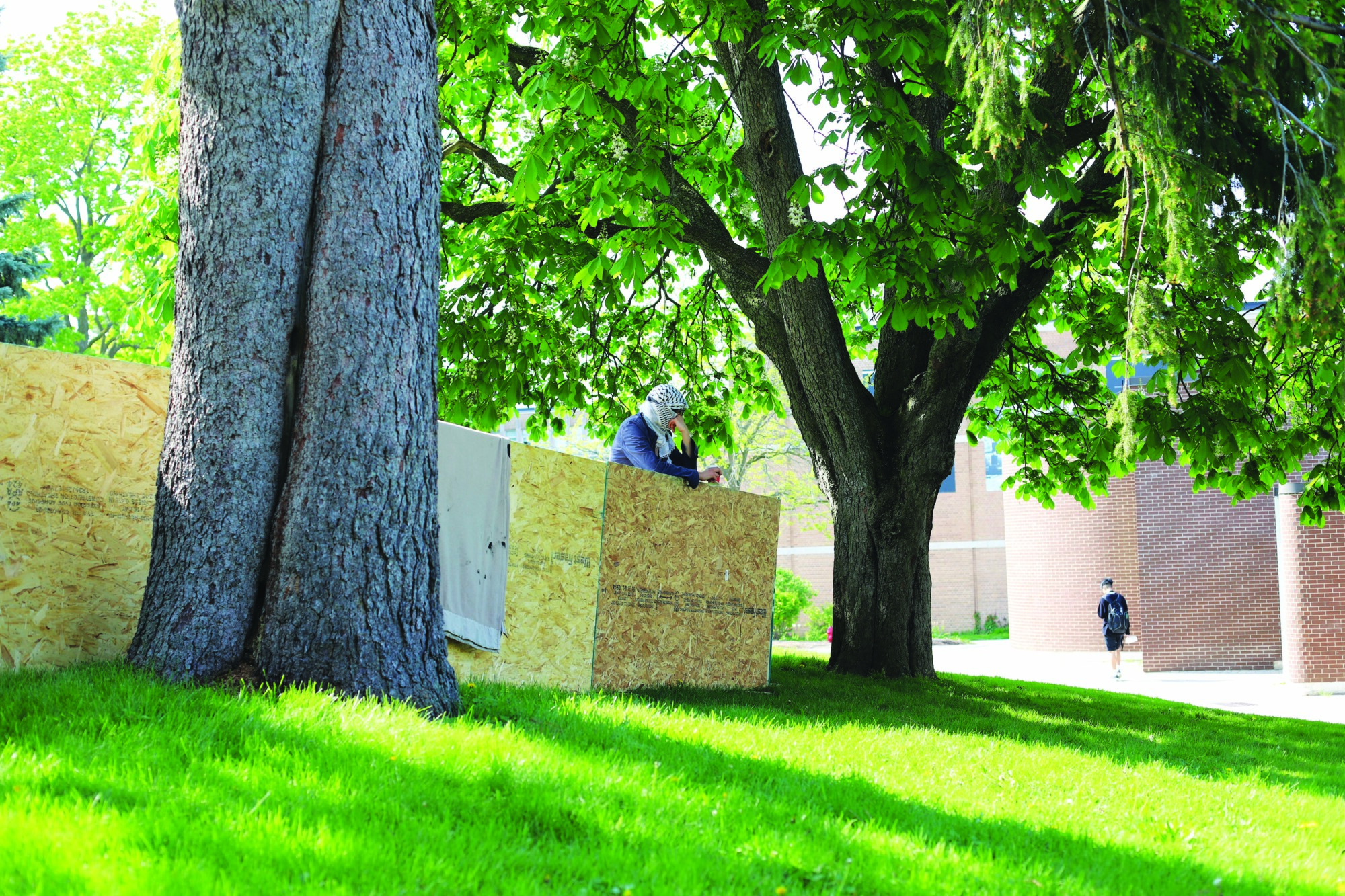 Photograph of a person on the UW campus, standing inside the encampment barricades while wearing a keffiyeh over their face and resting their head on their hand.