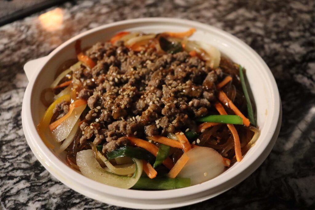Photo taken inside Taste of Seoul of a plate of Japchae topped with beef, onions, carrots, sesames seeds and sauce.