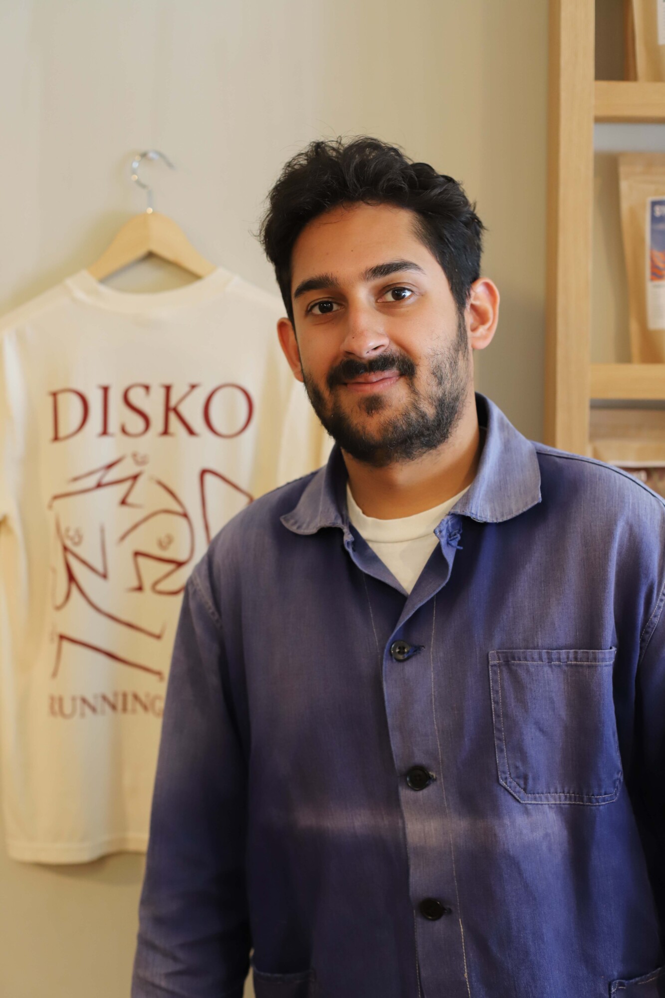 Photo of Arman Duggal, owner of Disko Coffee, posing inside his coffeeshop in front of a shirt with the name as the cafe on the front.