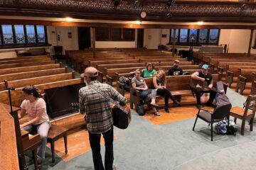 Photograph of a group of musicians sitting, talking and jamming together inside the Emmanuel United Church in Waterloo, Ontario.