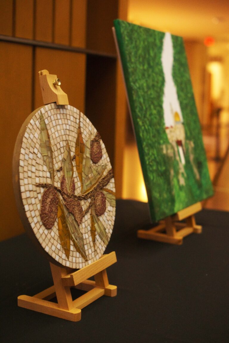 A visual arts display sitting on a table in the Kitchener City Hall. The piece in front is a round mosaic depicted an olive branch, and the piece in the back depicts a painting of a cityscape surrounded by leaves.
