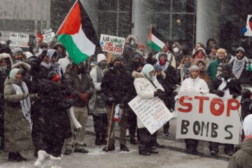 Protesters standing outside at Carl Zehr Square, Kitchener, waving Palestinian flags and holding signs reading "STOP BOMBS", "NO MONEY FOR WAR" and "CEASEFIRE". The protesters are wearing coats and it is snowing heavily.