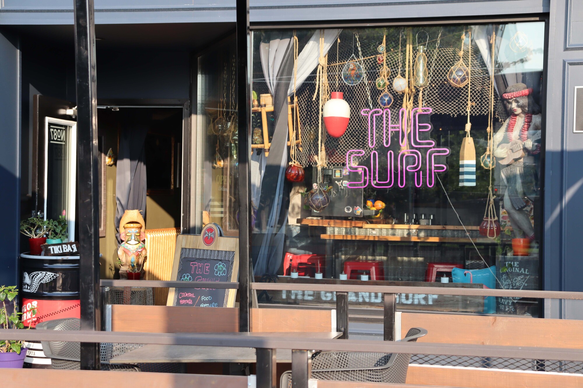 Photograph of the front entrance and window of The Grand Surf Lounge in Kitchener. The inside of the window is decorated with hung fishing nets, colourful glass lanterns and jars, a buoy, tiki figurines and a wooden mermaid playing the ukulele.