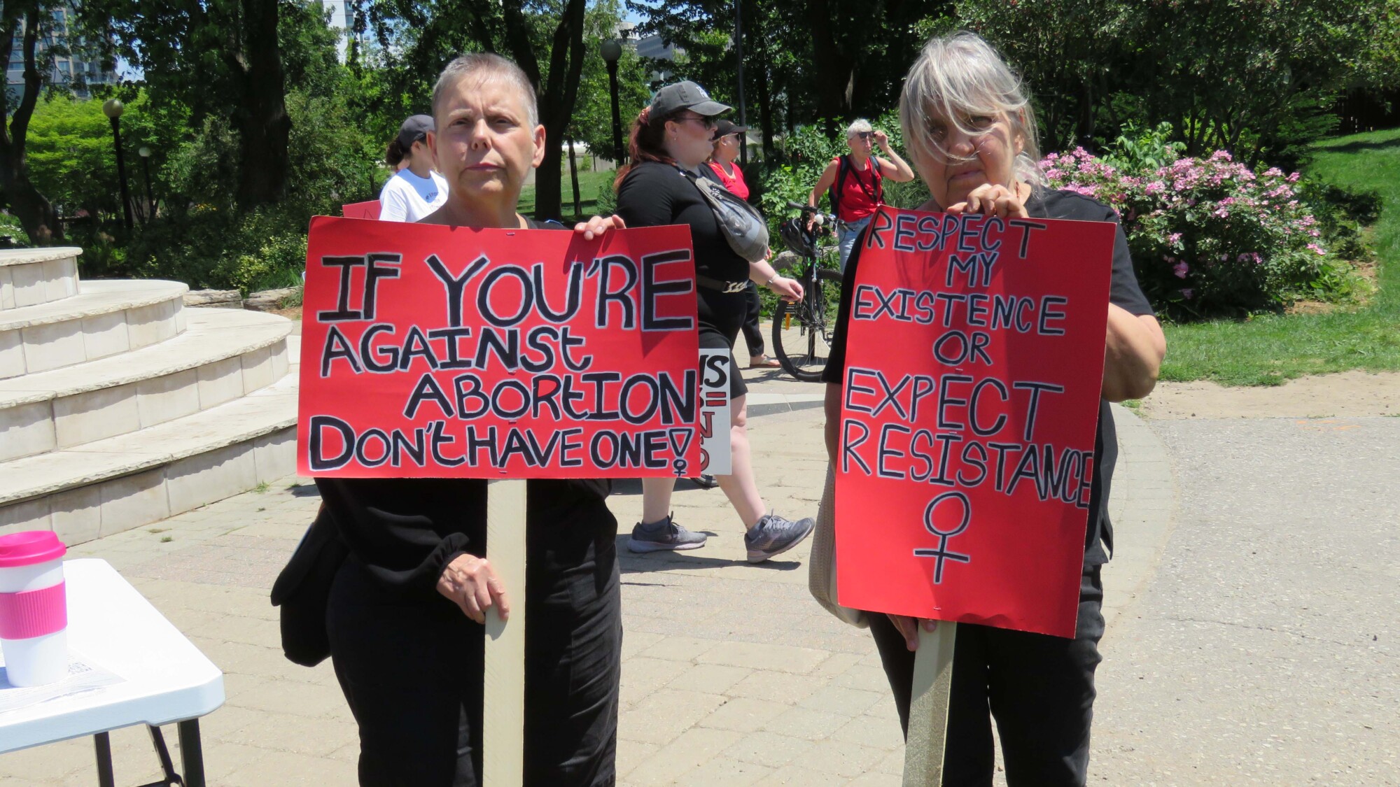 Photo of two protesters attending a rally held by the National Women's Strike Canada. The protesters are standing side by side in Willow River Park, Kitchener, holding up sighs reading "If you're against abortion, don't have one!" and "Respect my existence or expect resistance".