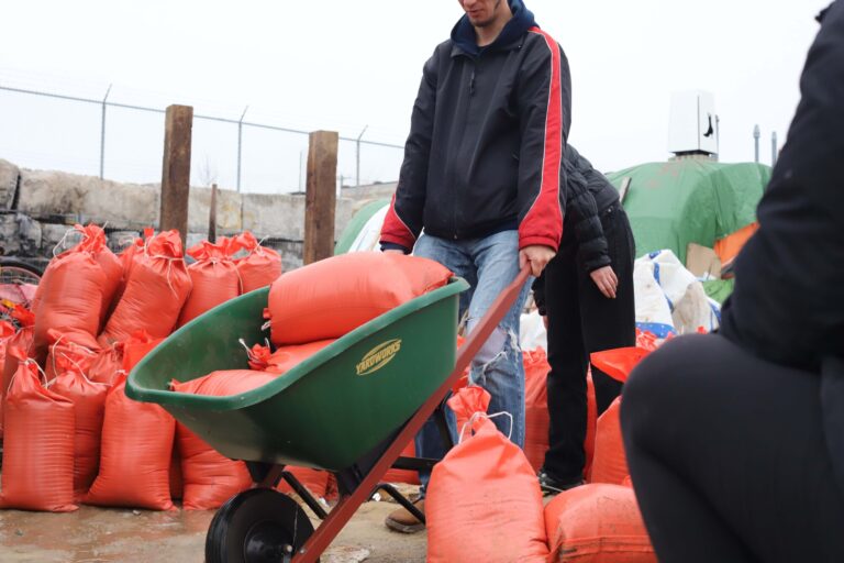 Photo of a person outside moving orange bags of sand in a wheelbarrow, while surrounded by similar bags filled with sand, as part of a volunteer effort to construct homes for Kitchener encampment residents.
