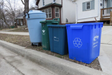 A garbage bin, a green bin and a blue box recycling bin is on a curb.