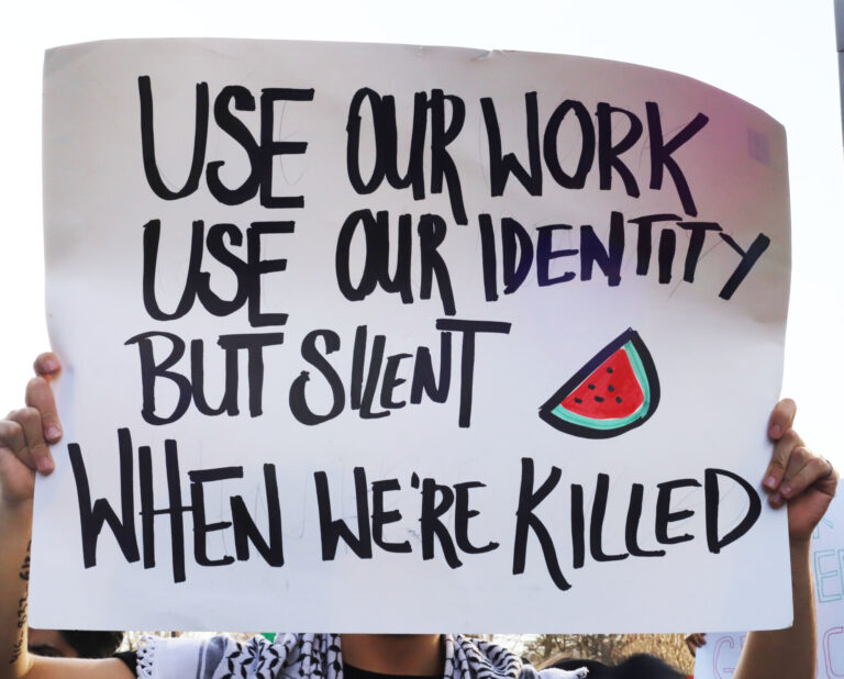 A person is holding up a sign in protest that reads "use our work, use our identity, but silent when we're killed"