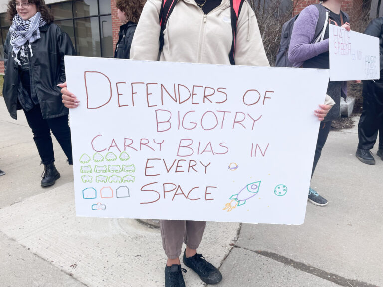 Photo of an anonymous protestor holding up a sign for the camera that reads "DEFENDERS OF BIGOTRY CARRY BIAS IN EVERY SPACE" and is decorated with fan art from the game Space Invaders.