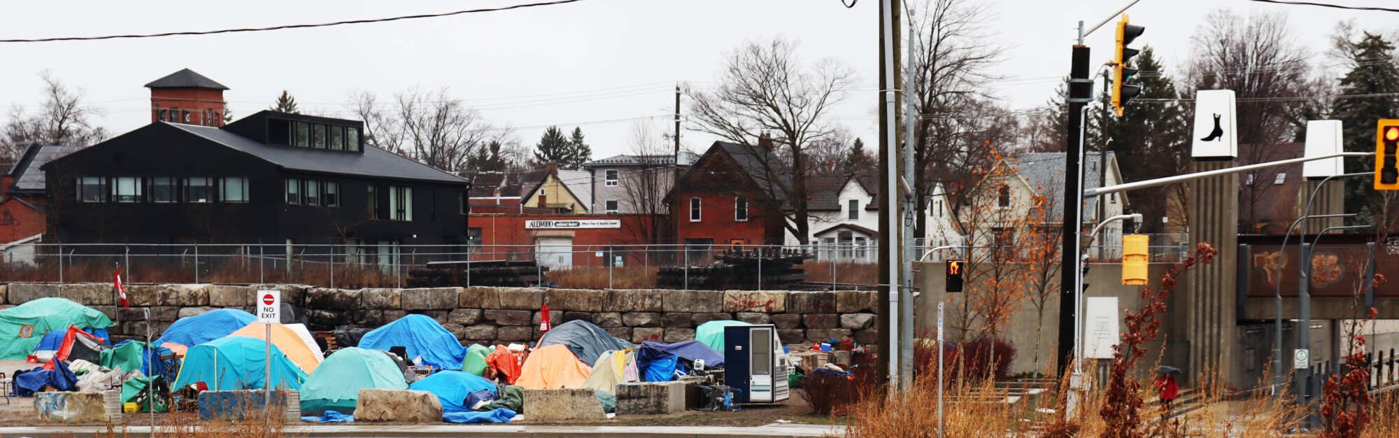 Photograph of the Victoria Street encampment in Kitchener, Ontario. A group of tents sit precariously together behind a traffic sign that reads "No Exit".