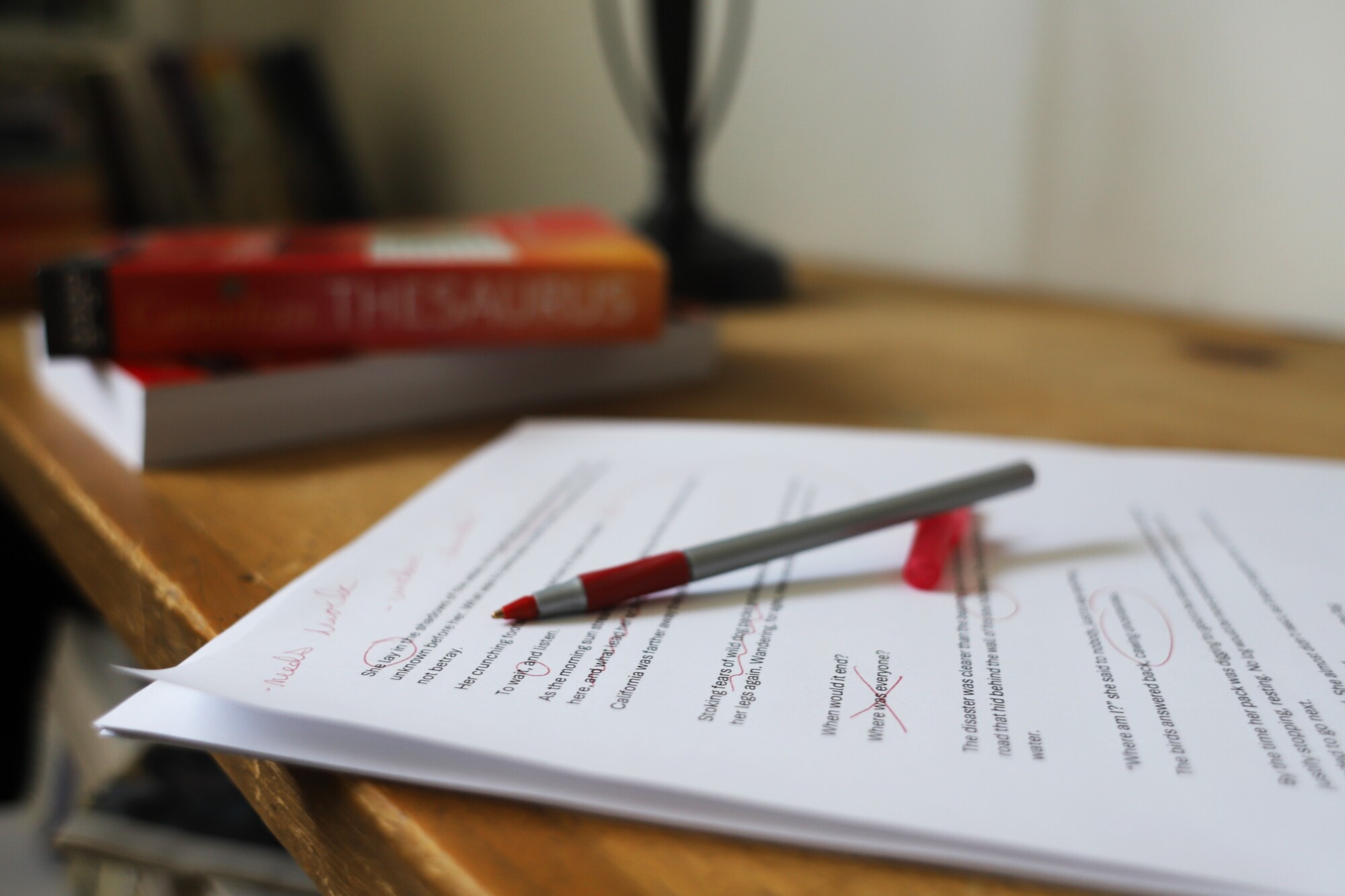 Photo of a short story that's been heavily marked up with a red pen. It's sitting inside on a desk beside a lamp and a thesaurus.