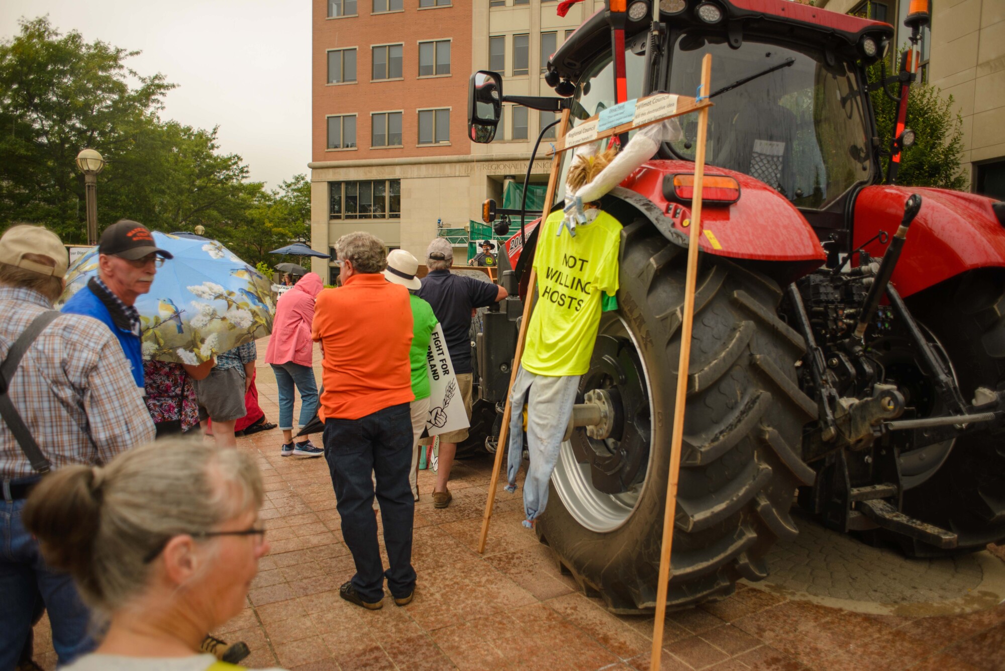 Photo of a scarecrow hoisted up on a giant tractor wheel, wearing a shirt that says "NOT WILLING HOSTS" at the Fight for Farmland rally held in Wilmot Township on August 28th, 2024.