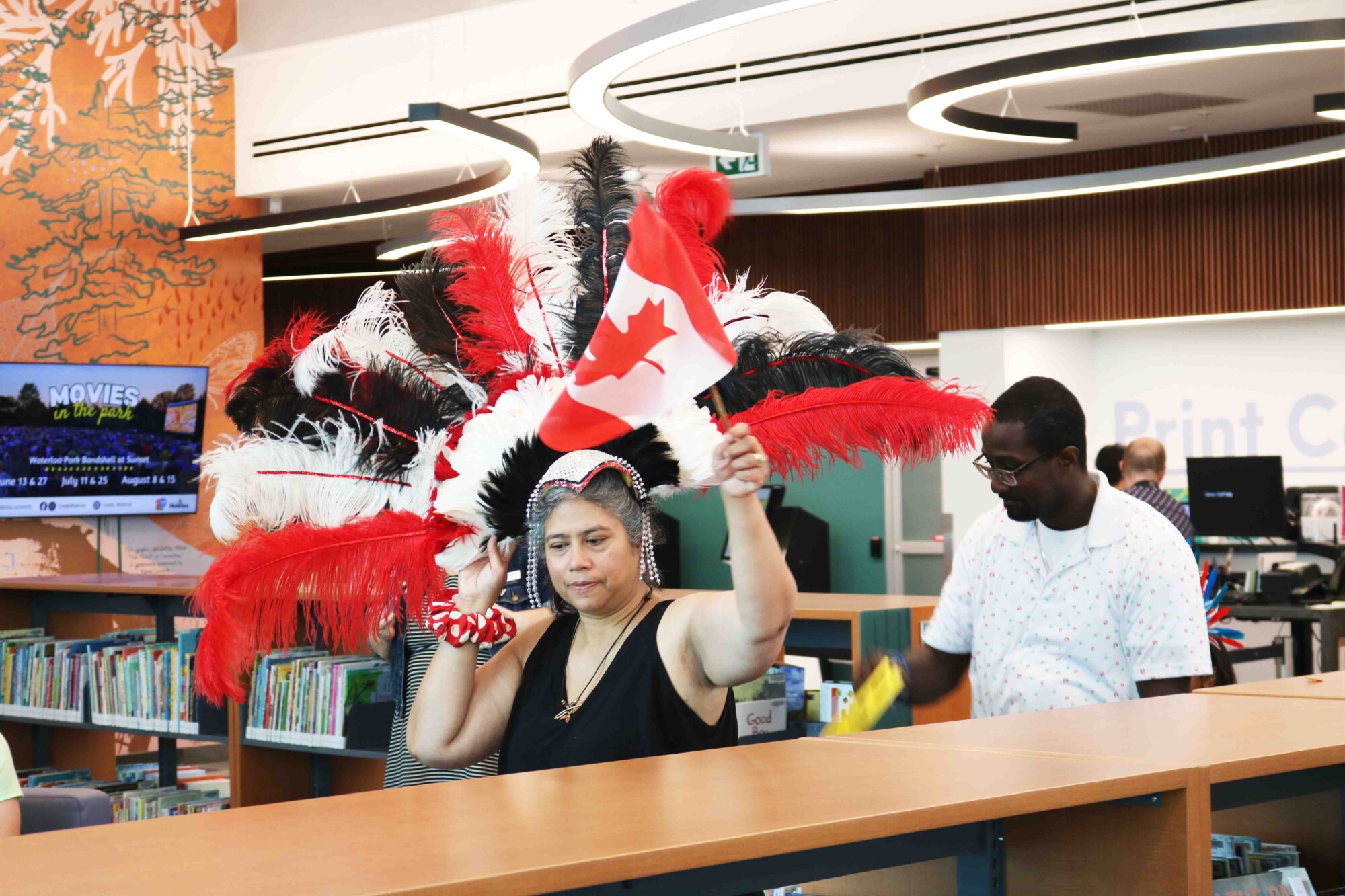 Photo of a woman wearing a colourful, feathered headpiece waving around a small Canadian flag as she parades around the Waterloo Public Library for attendees at the Waterloo Public Library Caribbean Carnival.