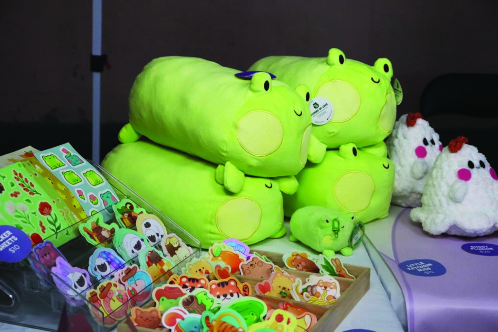 Photo of a pile of long frog plushies sitting on a vendor's table at the Kitchener Far East Asian Market, beside a display filled with colourful animal stickers and chickens made out of what appears to be yarn.