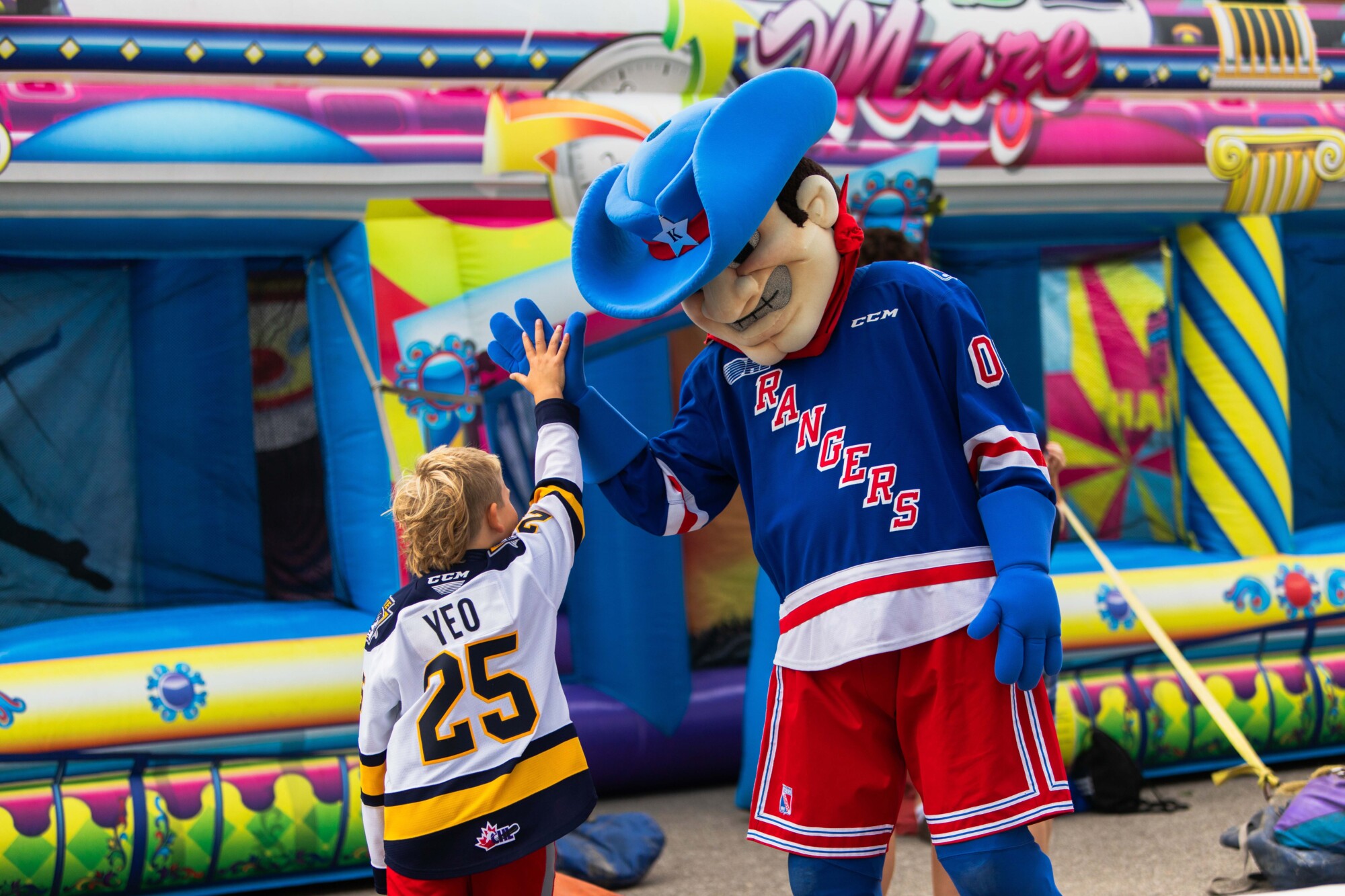 Photo of a child receiving a high five from a large costume of a white man with grey teeth at the Kitchener Ranger's preseason Fan Fest, held at the Kitchener Auditorium on September 1st, 2024.