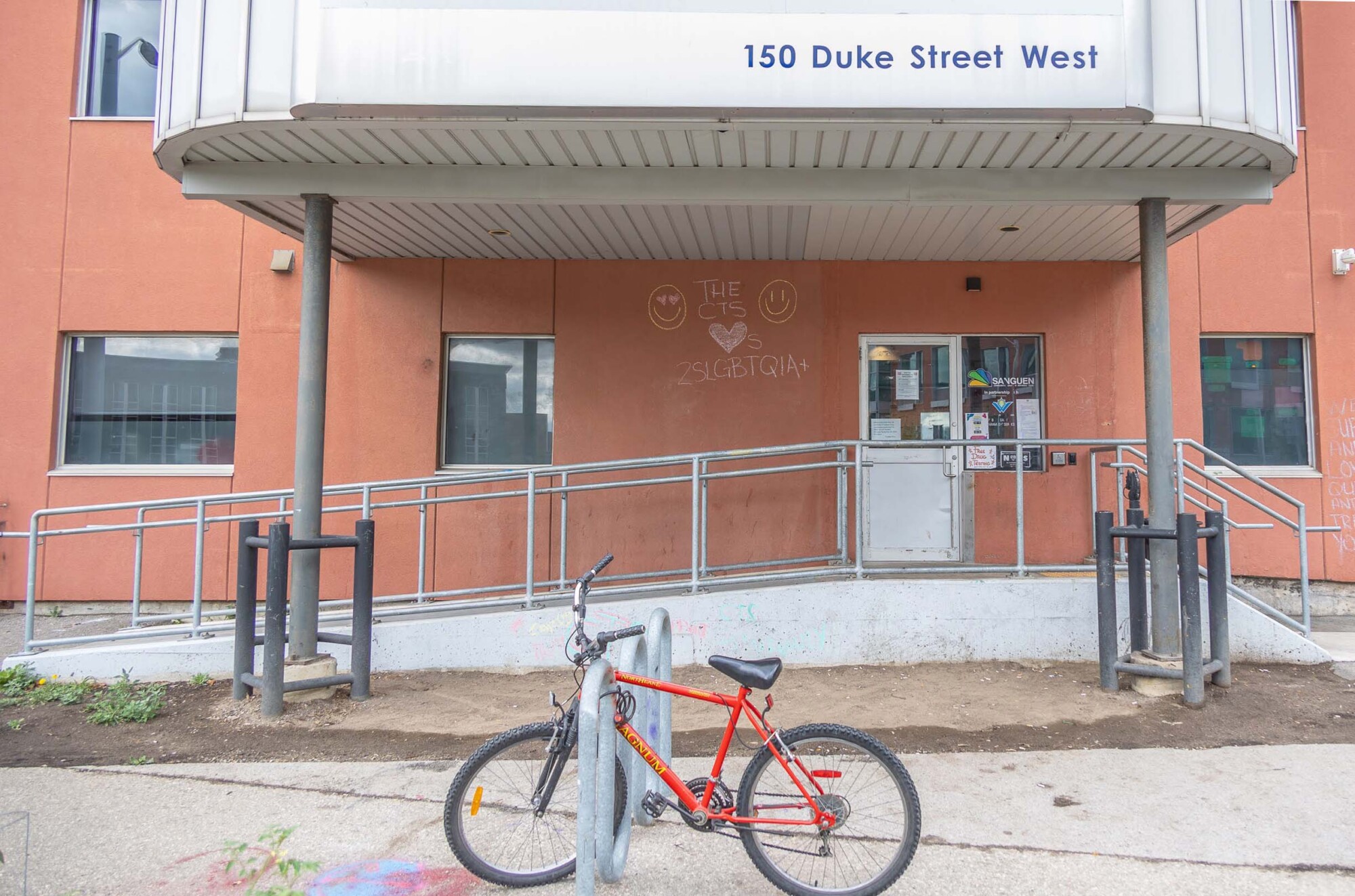 Photo of the front walkway of the Kitchener Consumption and Treatment Service building at 150 Duke Street, Kitchener. The building has a ramp, parking, and a yellow chemical waste container out front.