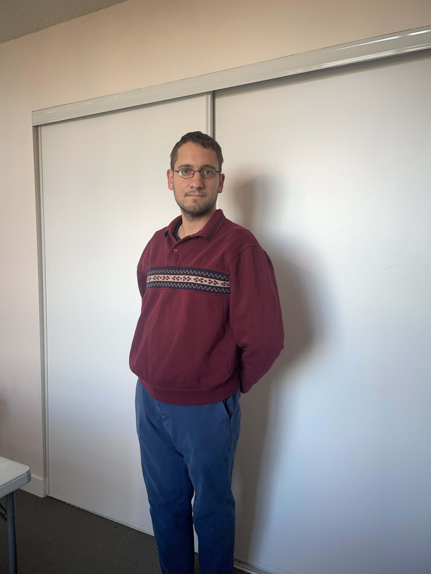 Photo of comedian Ben Stager standing in a beige room wearing a red sweater thinking about things as he looks at the camera.