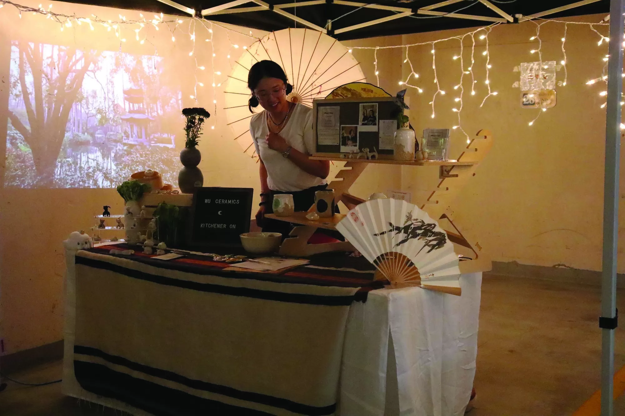 Photo of a vendor standing at her table, filled with merchandise, at the Far East Asian Market. There are strings of white fairy lights casting a soft glow over the fans, figurines and umbrellas she's selling. A fantastical outdoor scene is projected on the wall beside her.