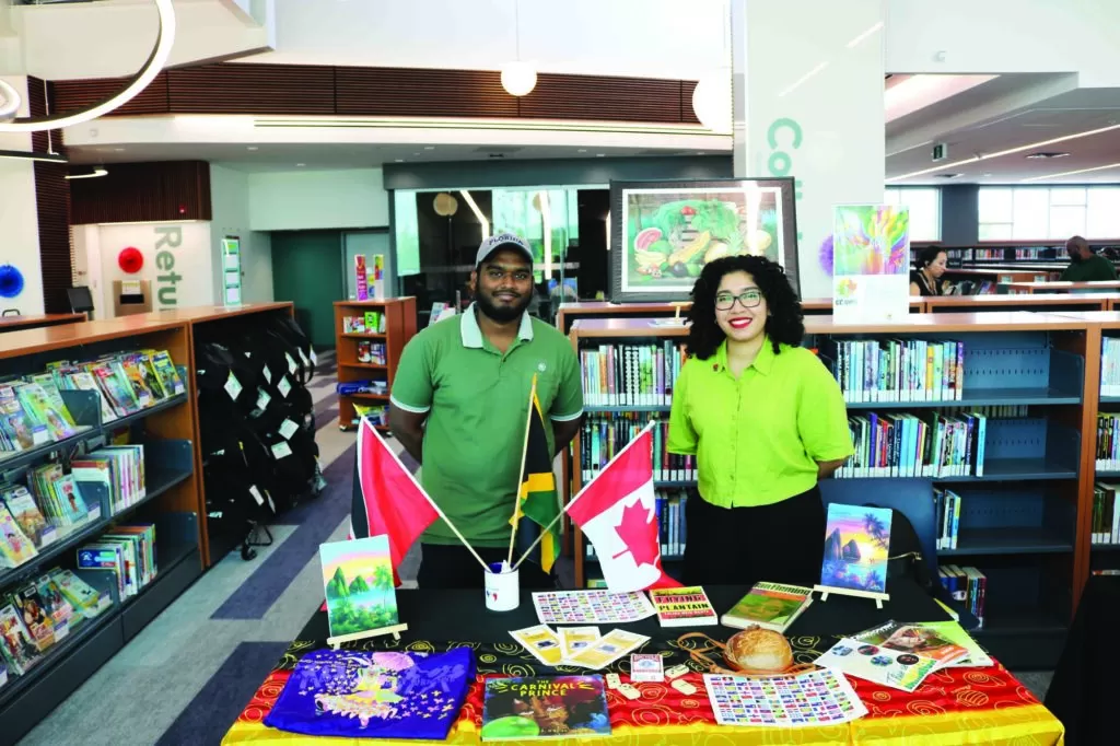 Photo of two people standing behind a table filled with stickers, books, and other educational materials during the Waterloo Public Library Caribbean Carnival.