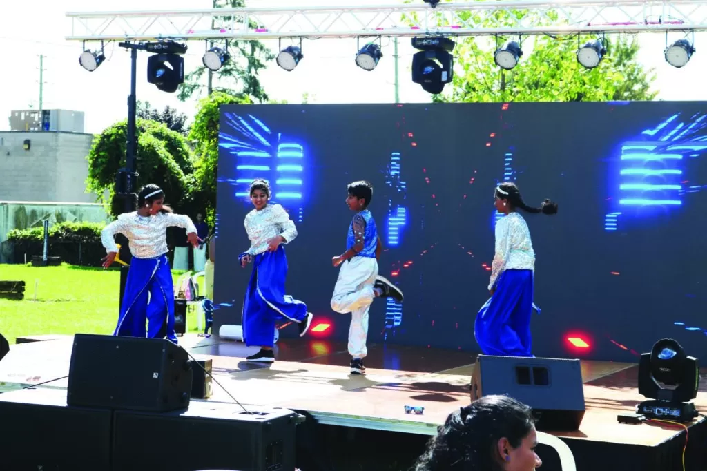 Photo of a team of young dancers performing on stage outside at the KW Onam Carnival. They're wearing sparkly blue and silver outfits and dancing in front of a dynamic, neon backdrop.